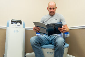 A patient in a t-shirt and jeans reads a magazine while he sits on the Emsella, urinary incontinence treatment near The Villages, FL.
