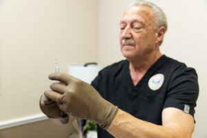 A provider with a mustache, wearing black scrubs and brown gloves, holds a syringe containing lip filler in Ocala.